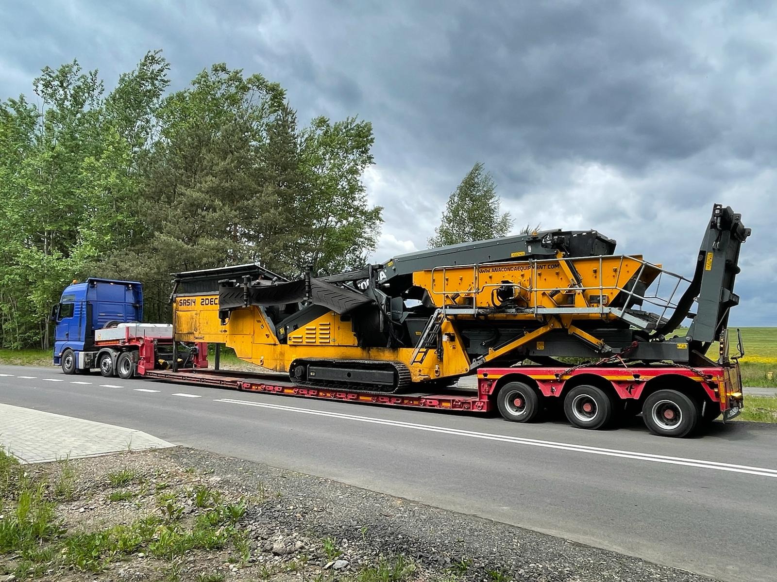 Niebieski ciągnik siodłowy z naczepą niskopodwoziową transportuje żółtą kruszarkę Anaconda SRS14 2DEC na tle pochmurnego nieba i zieleni.