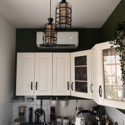 Everest - Biała klimatyzacja ścienna zamontowana nad rzędem białych szafek kuchennych na tle ściany w kolorze butelkowej zieleni, oświetlona przez dwie wiszące lampy w stylu industrialnym.