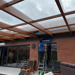 Everest - Mężczyzna na drabinie w niebieskiej koszulce, serwisujący klimatyzator zewnętrzny na tle ceglanej ściany i przeszklonego zadaszenia tarasu, z widocznymi narzędziami i odzyskanym płynem chłodniczym.