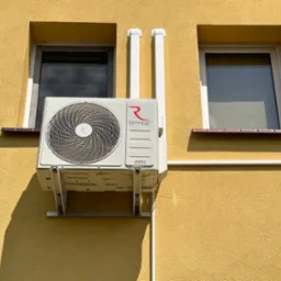 Biała jednostka zewnętrzna klimatyzatora Rotenso Hiro Series zamontowana na żółtej elewacji budynku między dwoma oknami, z widocznymi białymi rurami osłonowymi.