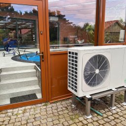 Everest - Biała pompa ciepła marki Seyra zamontowana na metalowej podstawie, widoczna na zewnątrz budynku z przeszkloną oranżerią i basenem, na brukowanej nawierzchni.