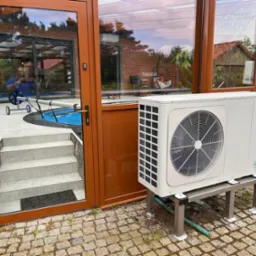 Biała pompa ciepła marki Seyra zamontowana na metalowej podstawie, widoczna na zewnątrz budynku z przeszkloną oranżerią i basenem, na brukowanej nawierzchni.