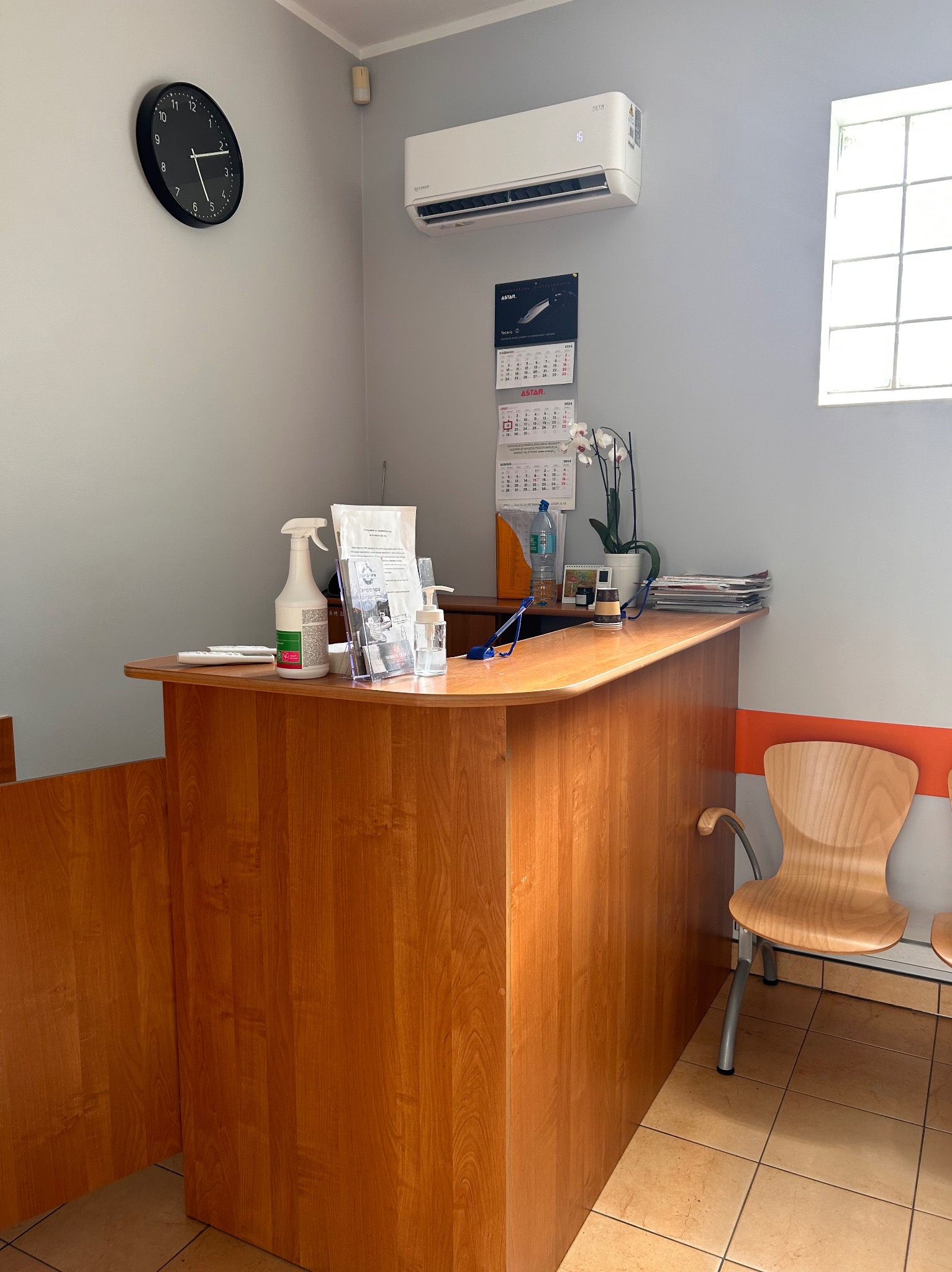 Recepcja biura z klimatyzatorem na ścianie, zegarem i kalendarzem z logo firmy ASTAR, na ladzie środki do dezynfekcji i ulotki informacyjne.