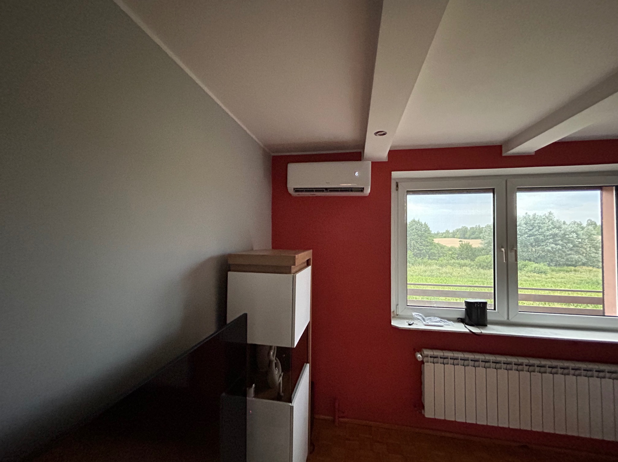 Biały klimatyzator ścienny zamontowany w pokoju z czerwoną ścianą, widok na zewnątrz przez okno z białymi ramami, grzejnik pod oknem, obok szafka RTV w tonacji bieli i drewna.