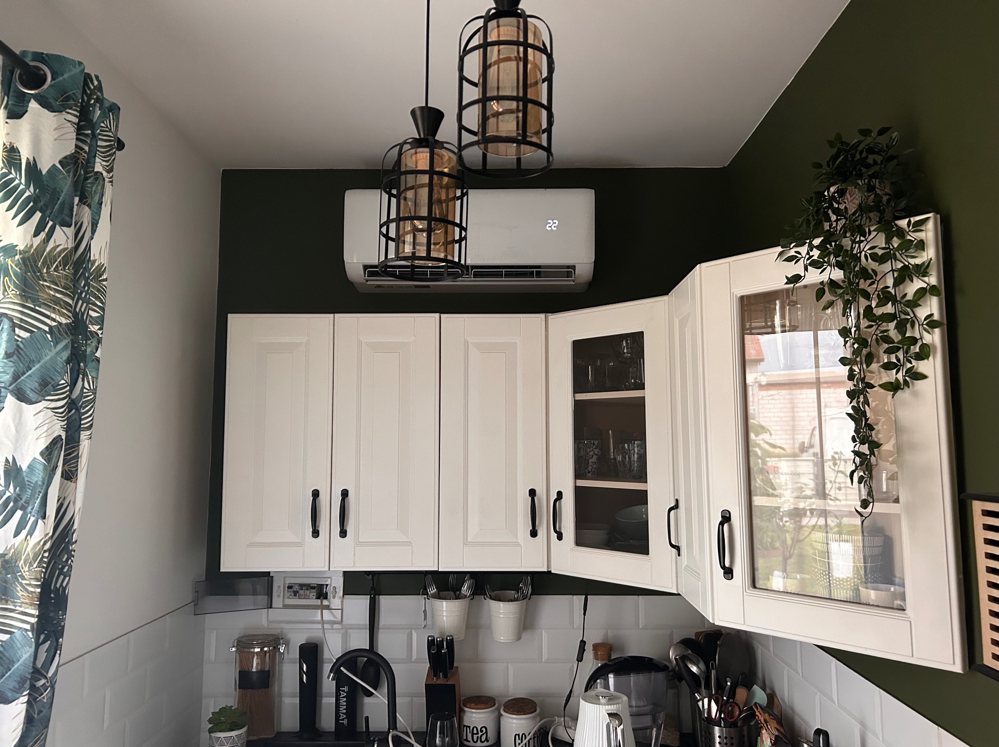 Biała klimatyzacja ścienna zamontowana nad rzędem białych szafek kuchennych na tle ściany w kolorze butelkowej zieleni, oświetlona przez dwie wiszące lampy w stylu industrialnym.