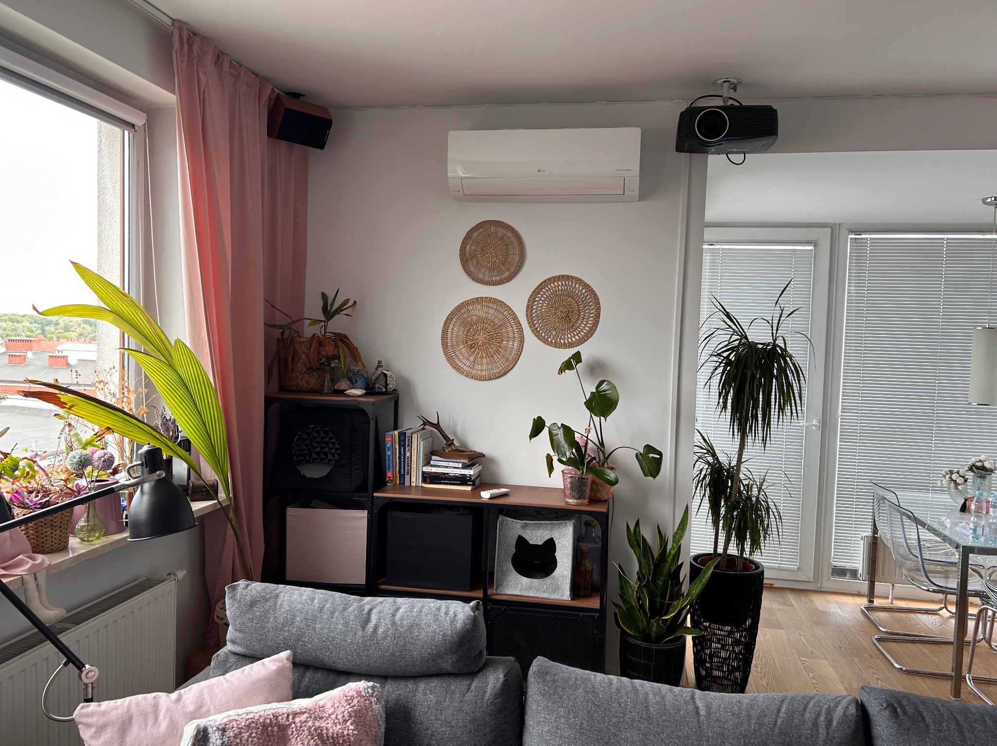 Wnętrze salonu z klimatyzatorem LG zamontowanym nad dekoracyjnymi, plecionymi talerzami na ścianie, widoczny projektor pod sufitem, regał z książkami i roślinami oraz szara sofa z poduszkami.