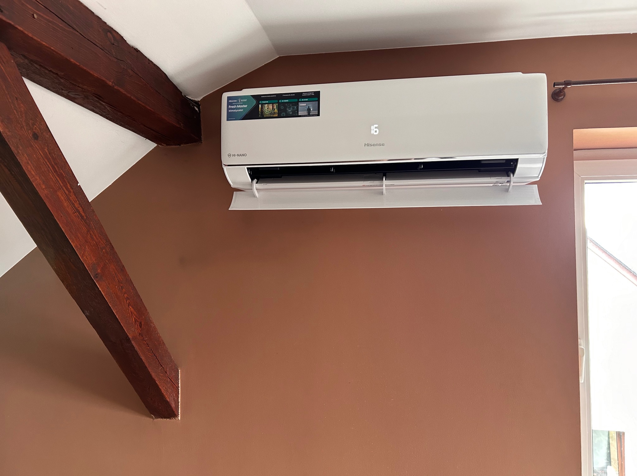 Biały klimatyzator Hisense zamontowany na brązowej ścianie w pomieszczeniu z drewnianą belką stropową i oknem w tle.