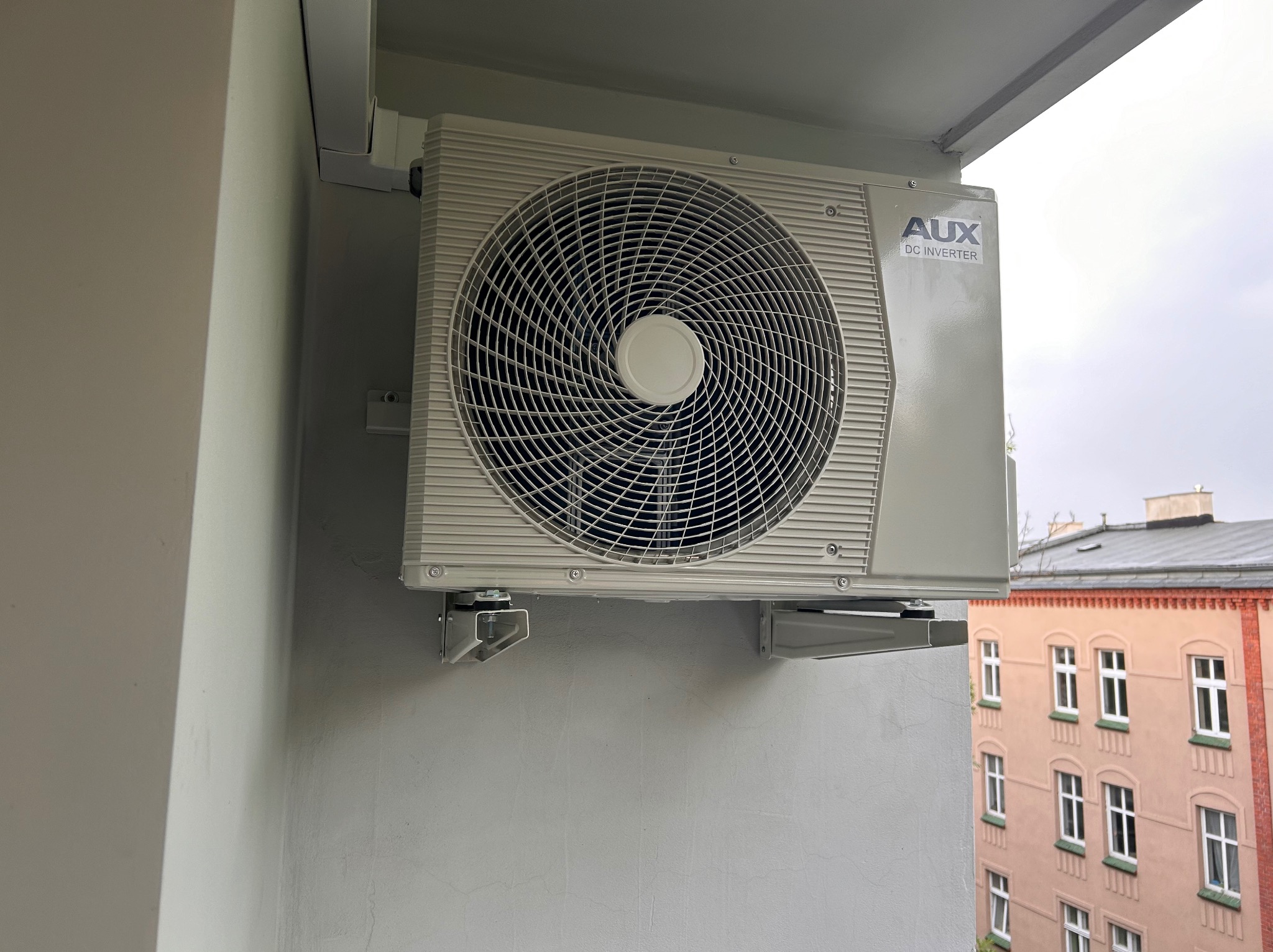 Zewnętrzna jednostka klimatyzacji AUX DC Inverter zamontowana na balkonie w bloku, widoczne mocowania i fragment budynku w tle.