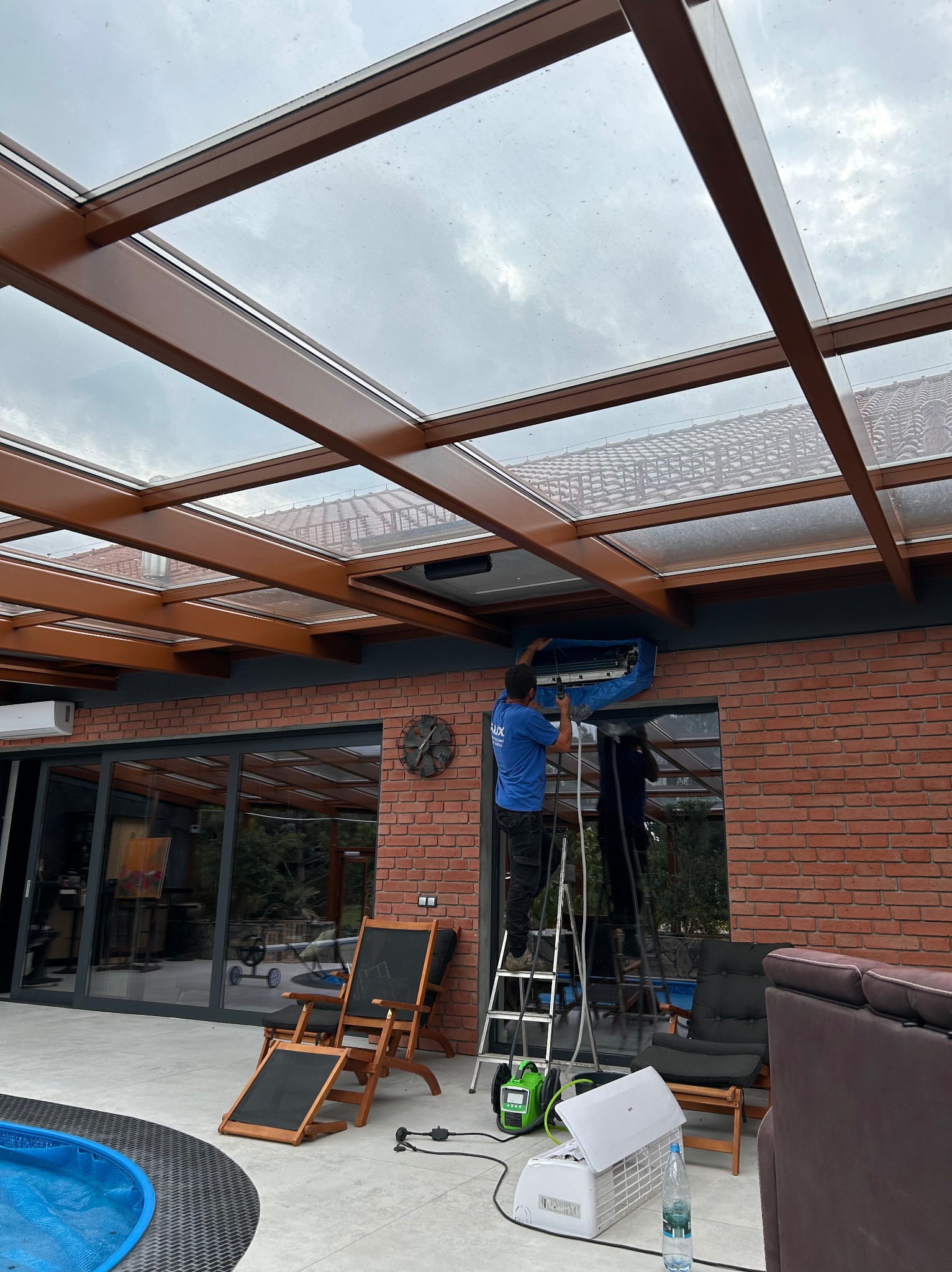 Mężczyzna na drabinie w niebieskiej koszulce, serwisujący klimatyzator zewnętrzny na tle ceglanej ściany i przeszklonego zadaszenia tarasu, z widocznymi narzędziami i odzyskanym płynem chłodniczym.
