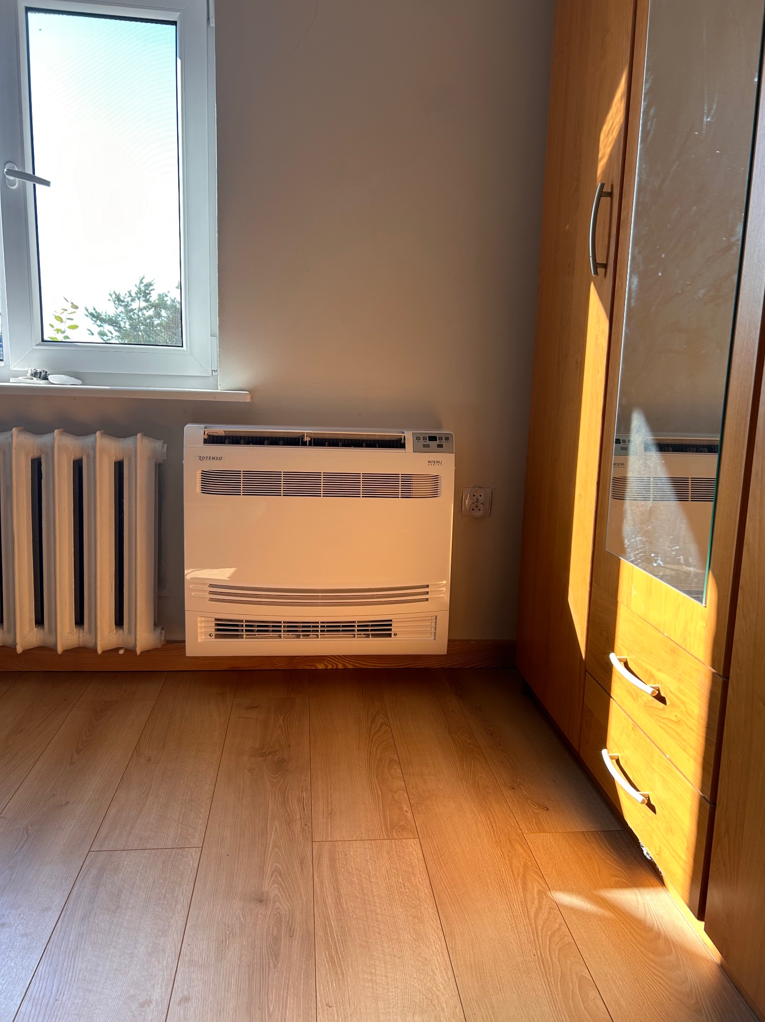 Stojący klimatyzator w pokoju, widoczny grzejnik żeberkowy, drewniana podłoga i szafa, oświetlone popołudniowym słońcem.