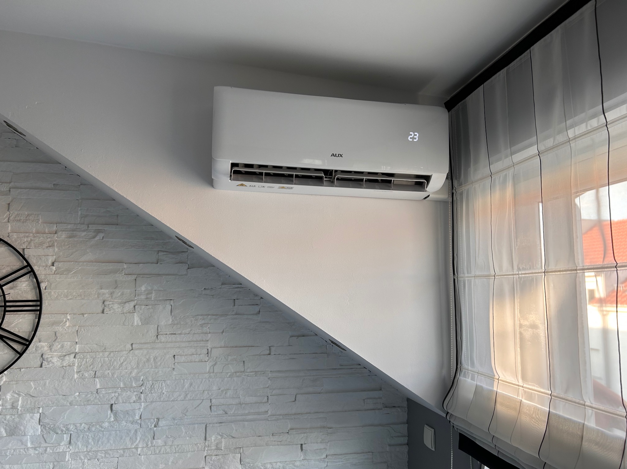 Biały klimatyzator marki AUX zamontowany na ścianie z dekoracyjną cegłą, wyświetlający temperaturę 23 stopnie Celsjusza, obok okna z roletami rzymskimi.
