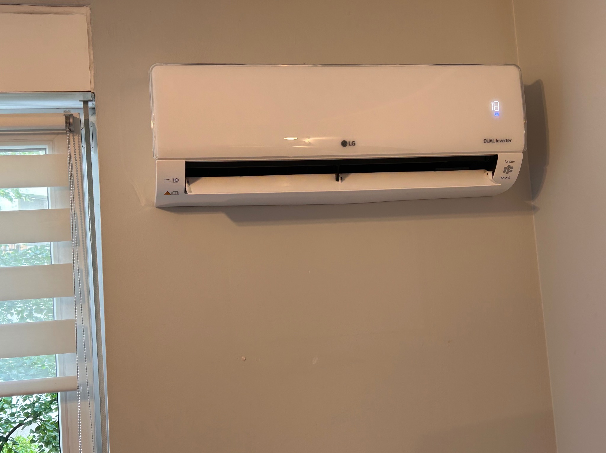 Biały klimatyzator LG Dual Inverter zamontowany na beżowej ścianie, widoczny wyświetlacz temperatury ustawiony na 18 stopni, żaluzje okienne w tle.