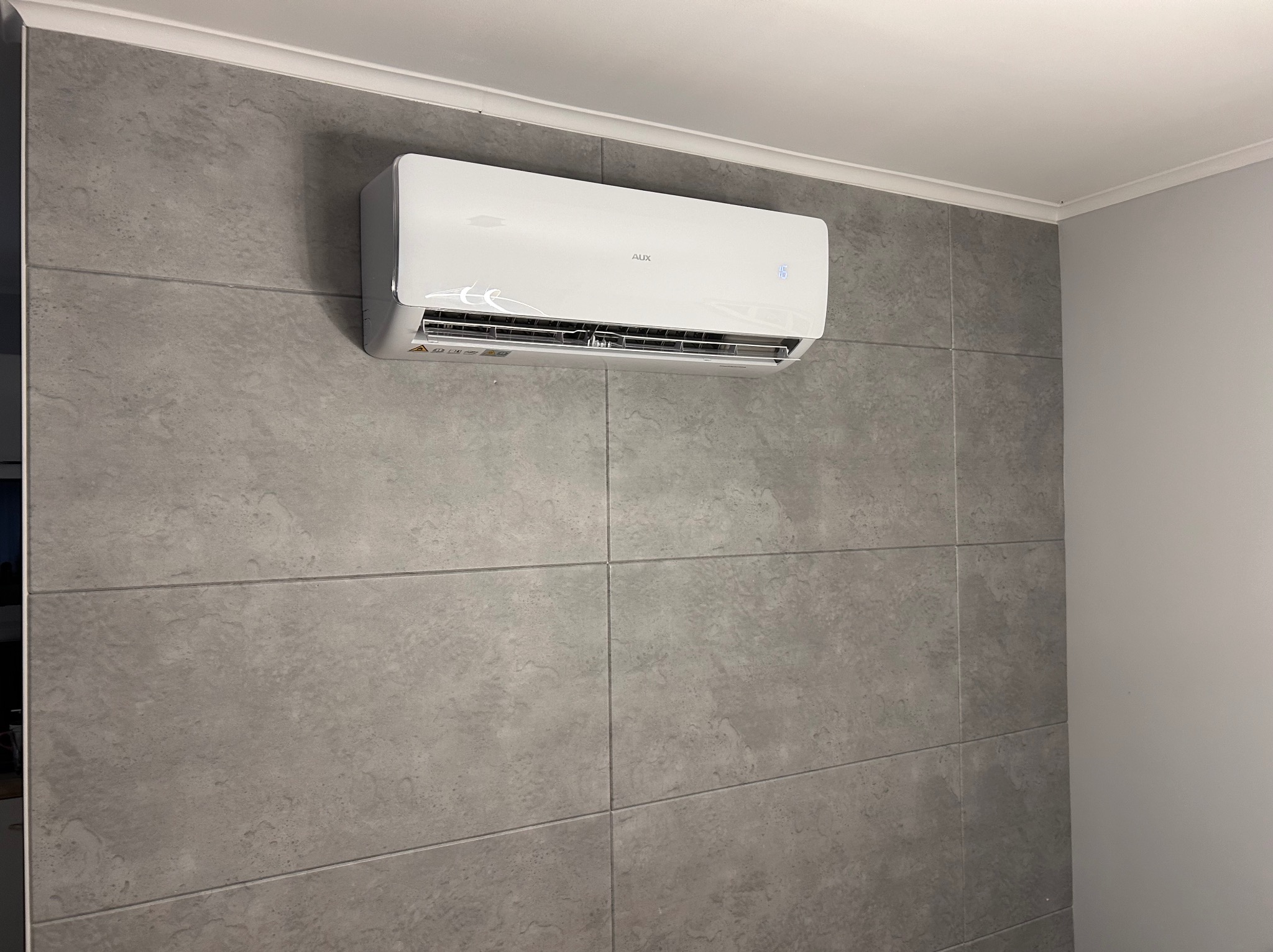 Biały klimatyzator AUX zamontowany na szarej ścianie z płytek imitujących kamień, widoczny wyświetlacz temperatury.