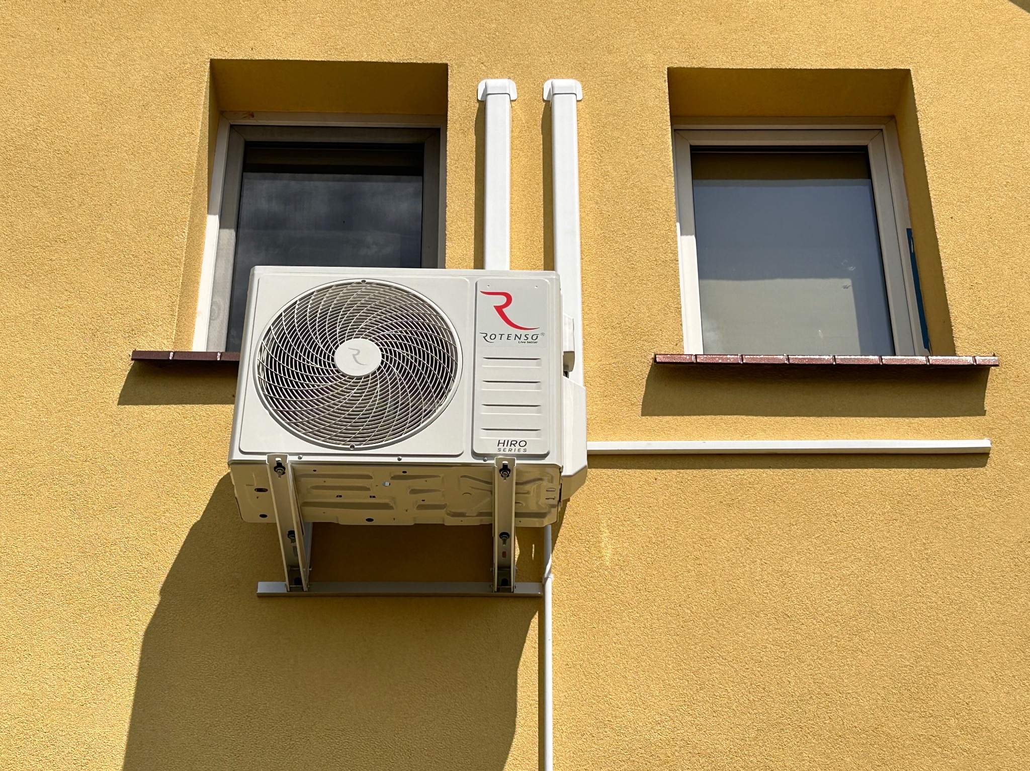 Biała jednostka zewnętrzna klimatyzatora Rotenso Hiro Series zamontowana na żółtej elewacji budynku między dwoma oknami, z widocznymi białymi rurami osłonowymi.