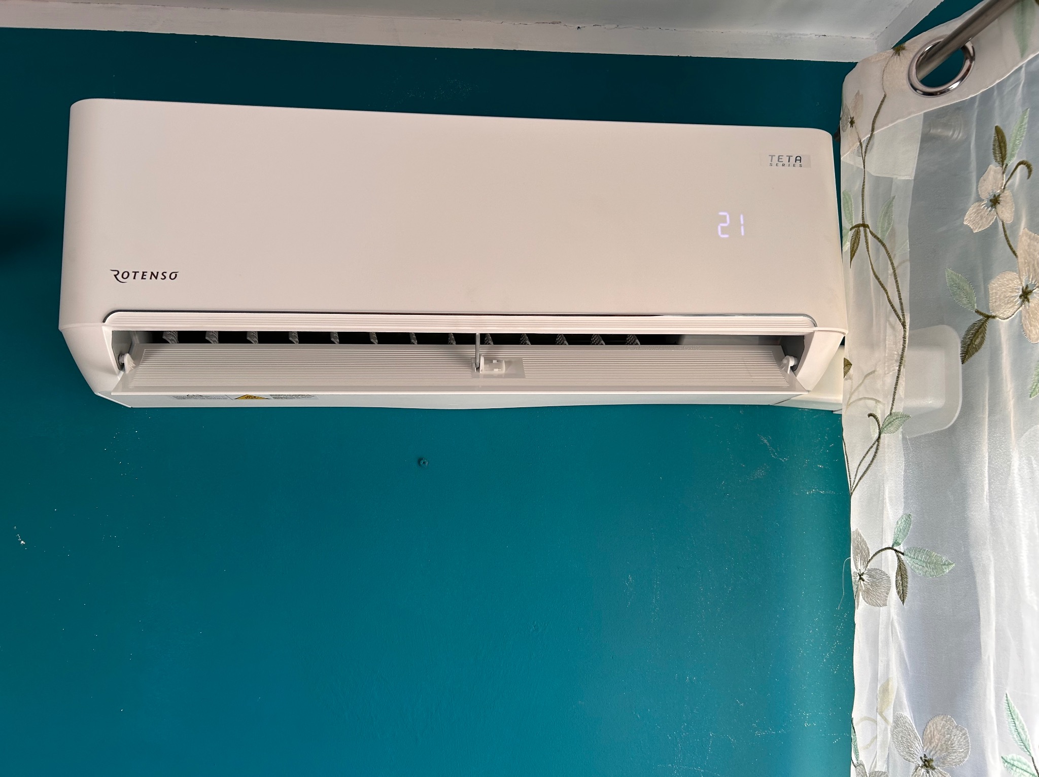 Biały klimatyzator Rotenso Teta Series zamontowany na turkusowej ścianie, widoczny wyświetlacz z temperaturą 21 stopni Celsjusza, obok zwiewna firanka w kwiaty.