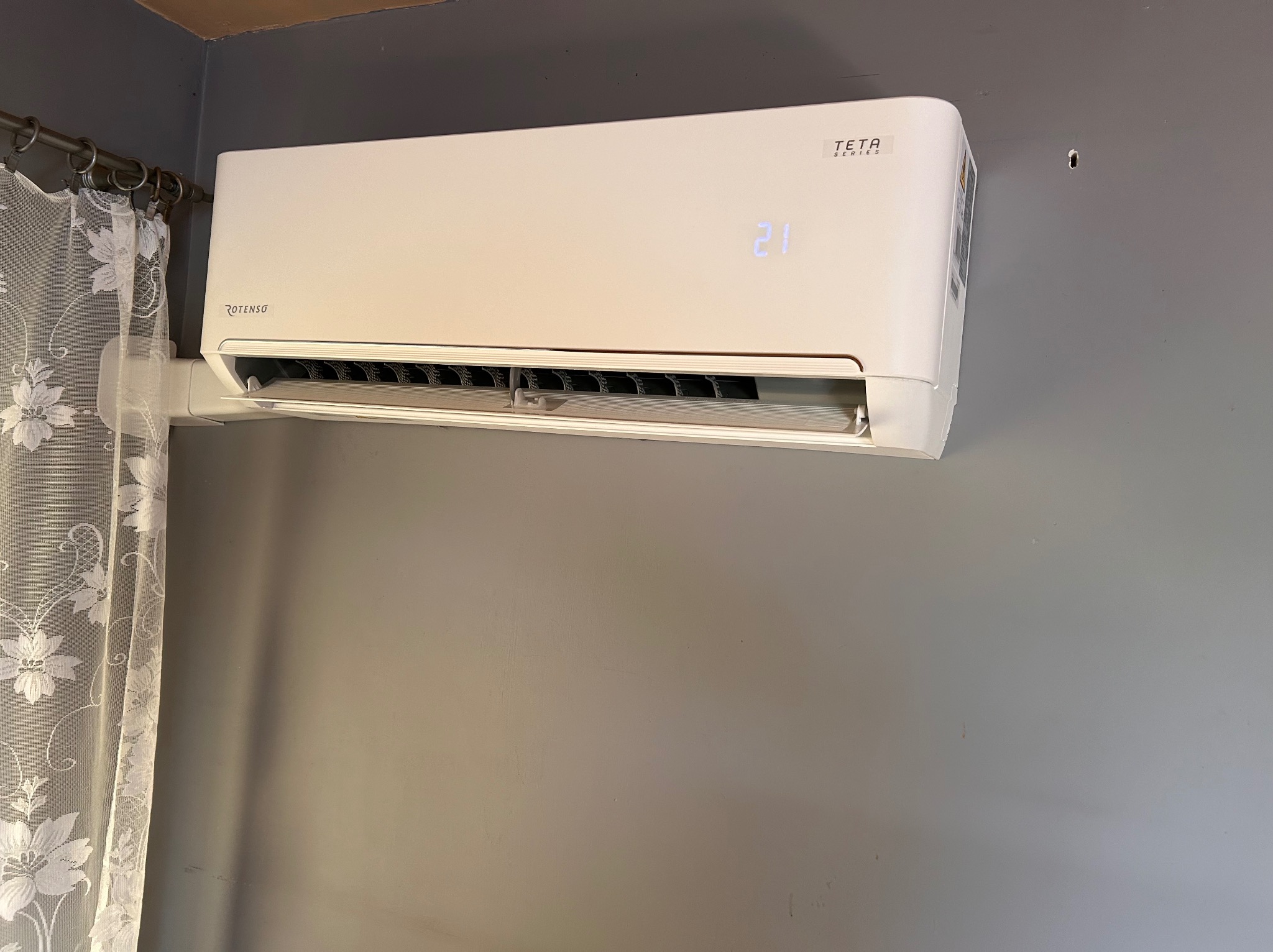 Biały klimatyzator Rotenso Teta Series zamontowany na szarej ścianie, z wyświetlaczem pokazującym temperaturę 21 stopni, obok firanki w kwiaty.