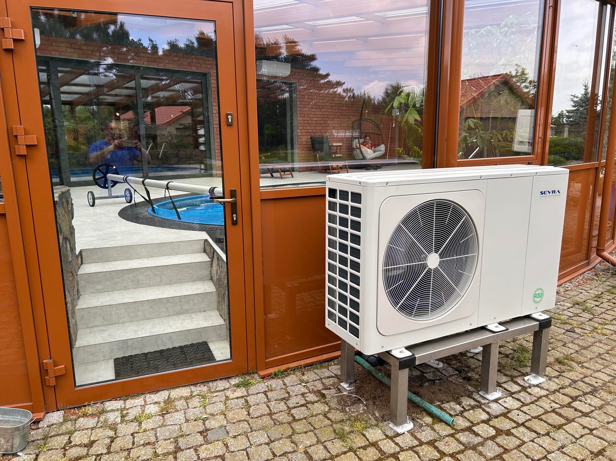 Biała pompa ciepła marki Seyra zamontowana na metalowej podstawie, widoczna na zewnątrz budynku z przeszkloną oranżerią i basenem, na brukowanej nawierzchni.