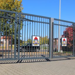 DEMEX SPÓŁKA Z OGRANICZONĄ ODPOWIEDZIALNOŚCIĄ - Szara, metalowa brama wjazdowa z automatyką, z czterema tablicami z logo Państwowej Straży Pożarnej, na tle jesiennych drzew i chodnika z kostki brukowej.