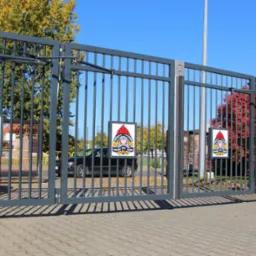 Szara, metalowa brama wjazdowa z automatyką, z czterema tablicami z logo Państwowej Straży Pożarnej, na tle jesiennych drzew i chodnika z kostki brukowej.