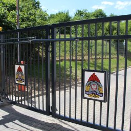 DEMEX SPÓŁKA Z OGRANICZONĄ ODPOWIEDZIALNOŚCIĄ - Czarna brama wjazdowa z automatyką i tabliczką Państwowej Straży Pożarnej na tle zieleni i kostki brukowej.