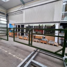 DEMEX SPÓŁKA Z OGRANICZONĄ ODPOWIEDZIALNOŚCIĄ - Zielona, metalowa brama z siatki, częściowo otwarta, zamontowana na metalowej konstrukcji w hali produkcyjnej, z widocznymi paletami i skrzyniami w tle.