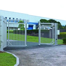 DEMEX SPÓŁKA Z OGRANICZONĄ ODPOWIEDZIALNOŚCIĄ - Otwarta, metalowa brama przemysłowa w kolorze szarym, z widocznym budynkiem przemysłowym w tle i zadbaną zielenią wokół.