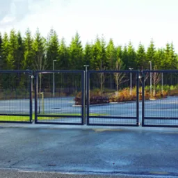 Czarna, czteroskrzydłowa brama przemysłowa z automatyką i panelem numerycznym, zamontowana na tle ogrodzenia z siatki i rzędu drzew iglastych.