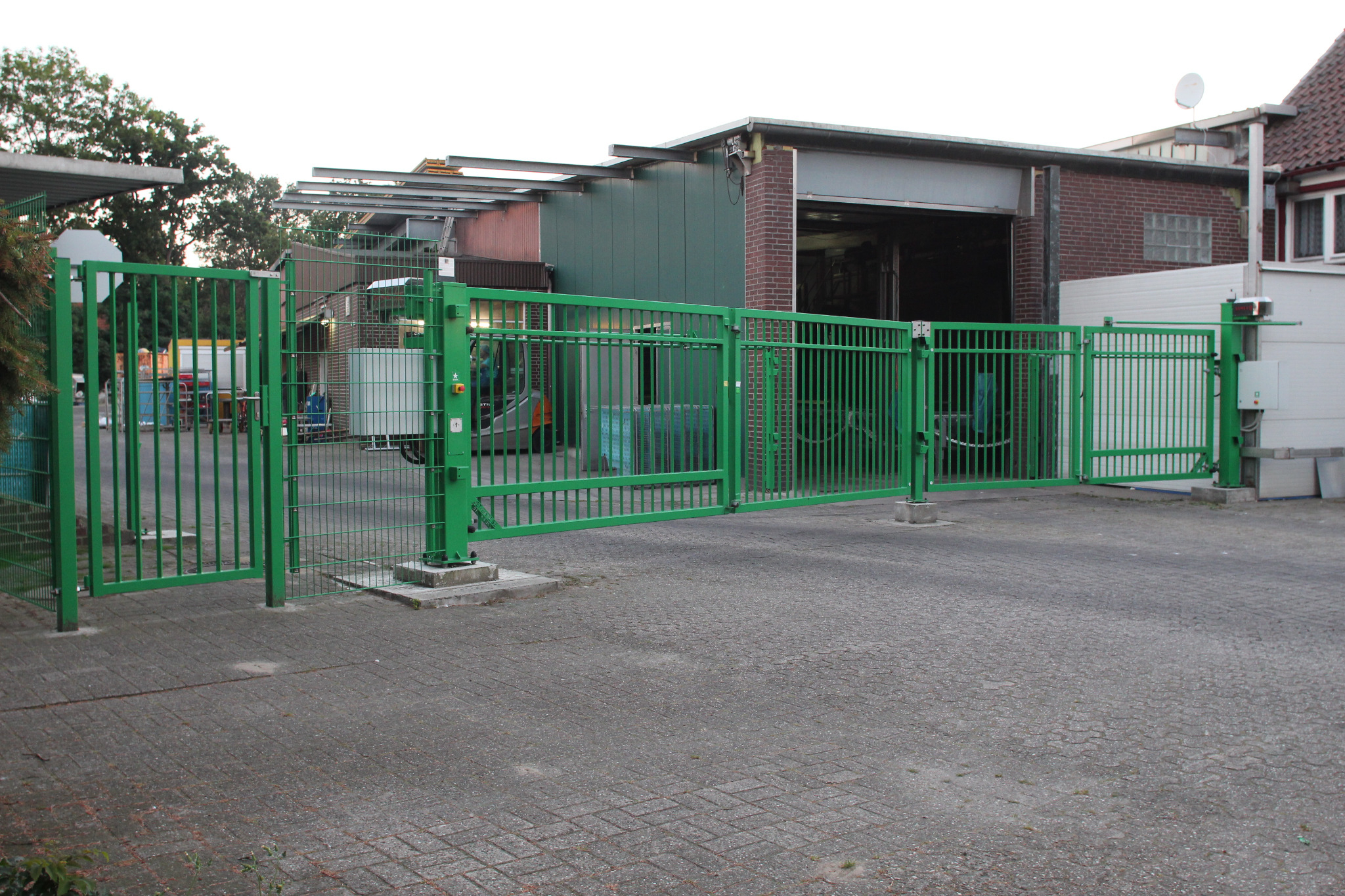 Zielona, metalowa brama przemysłowa z automatyką na tle budynku magazynowego z ceglaną elewacją i betonowym podjazdem.