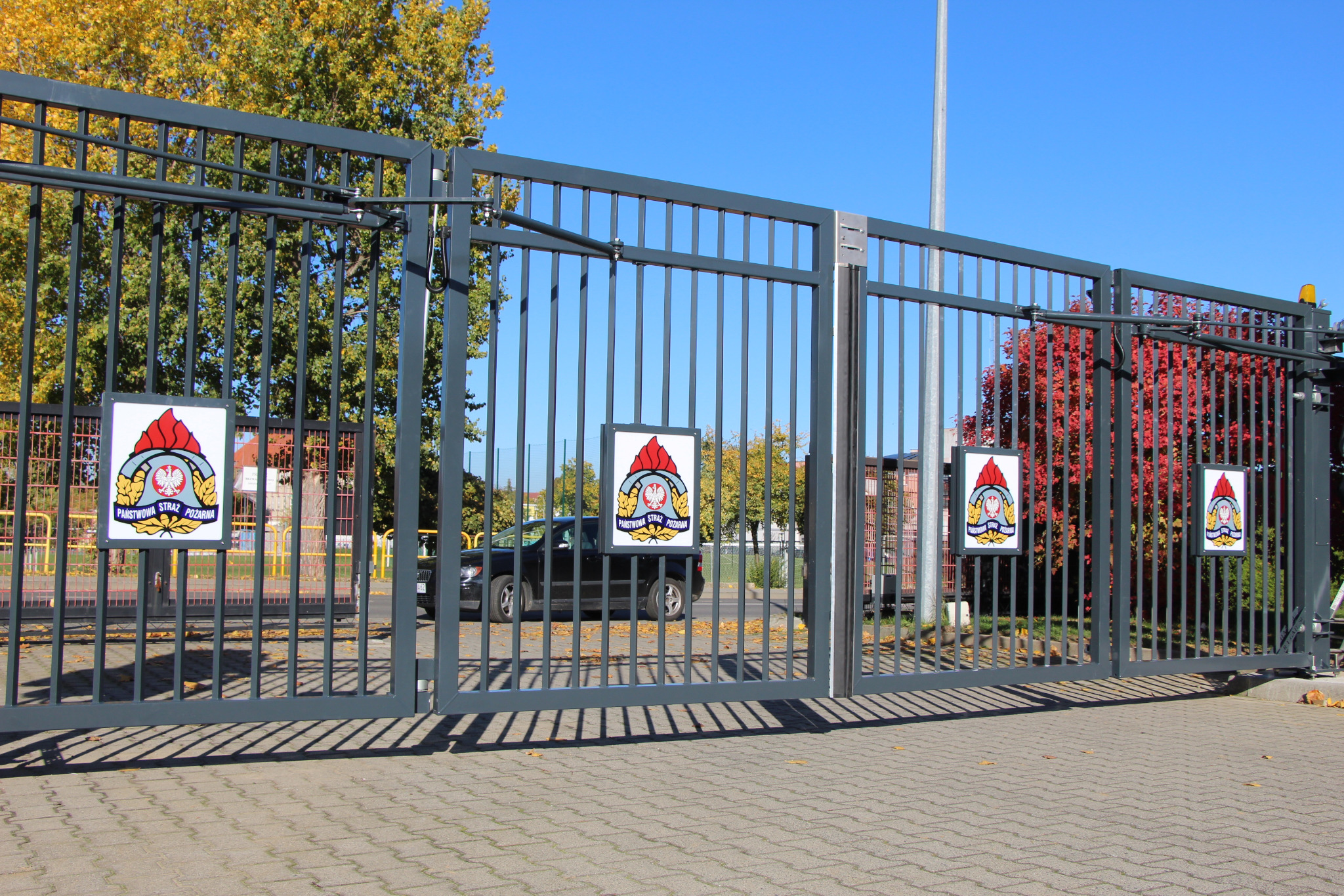 Szara, metalowa brama wjazdowa z automatyką, z czterema tablicami z logo Państwowej Straży Pożarnej, na tle jesiennych drzew i chodnika z kostki brukowej.