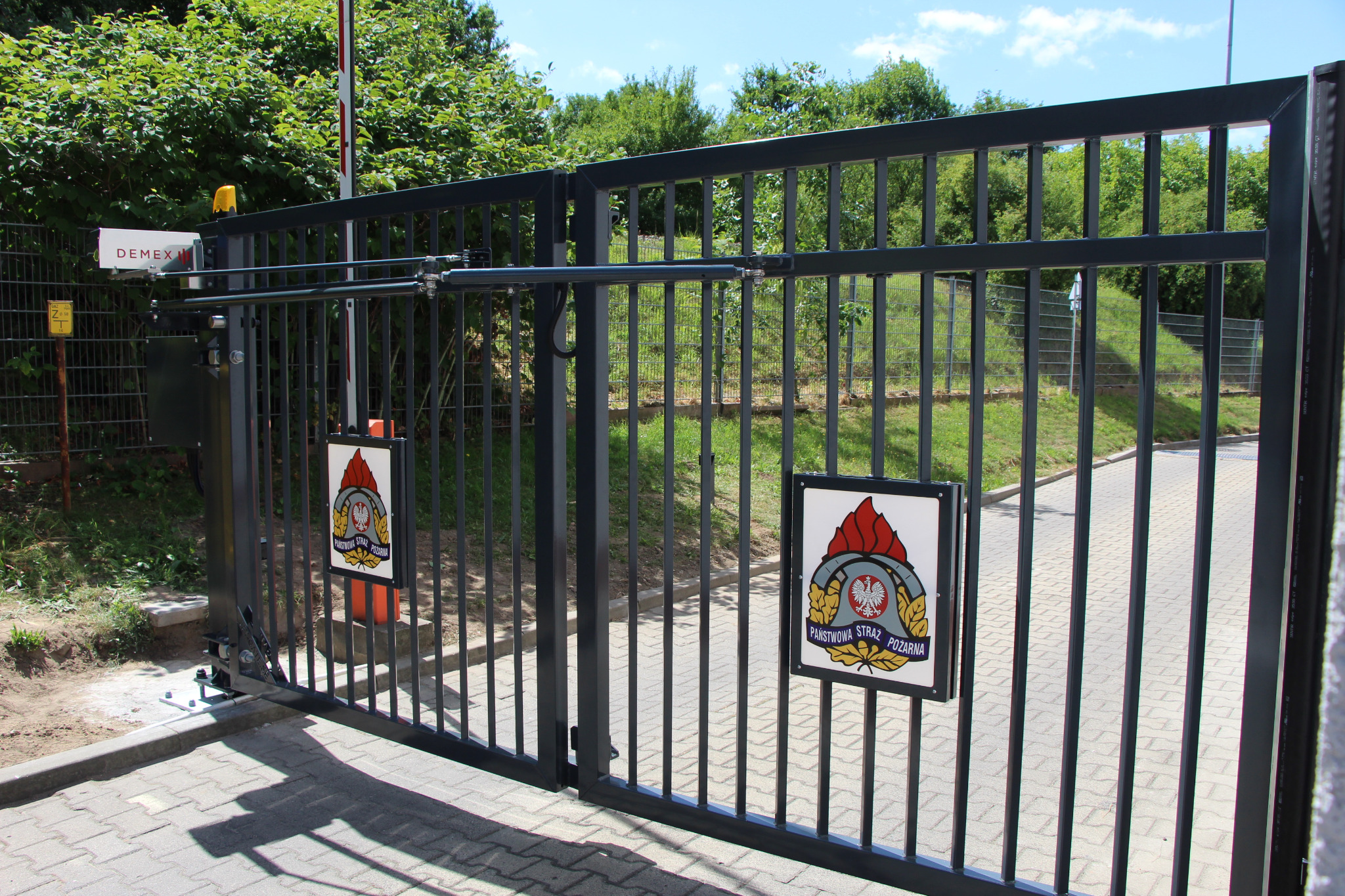 Czarna brama wjazdowa z automatyką i tabliczką Państwowej Straży Pożarnej na tle zieleni i kostki brukowej.