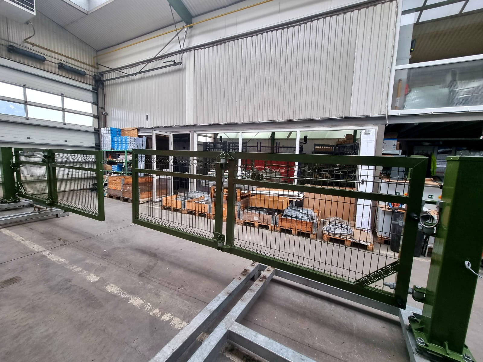 Zielona, metalowa brama z siatki, częściowo otwarta, zamontowana na metalowej konstrukcji w hali produkcyjnej, z widocznymi paletami i skrzyniami w tle.
