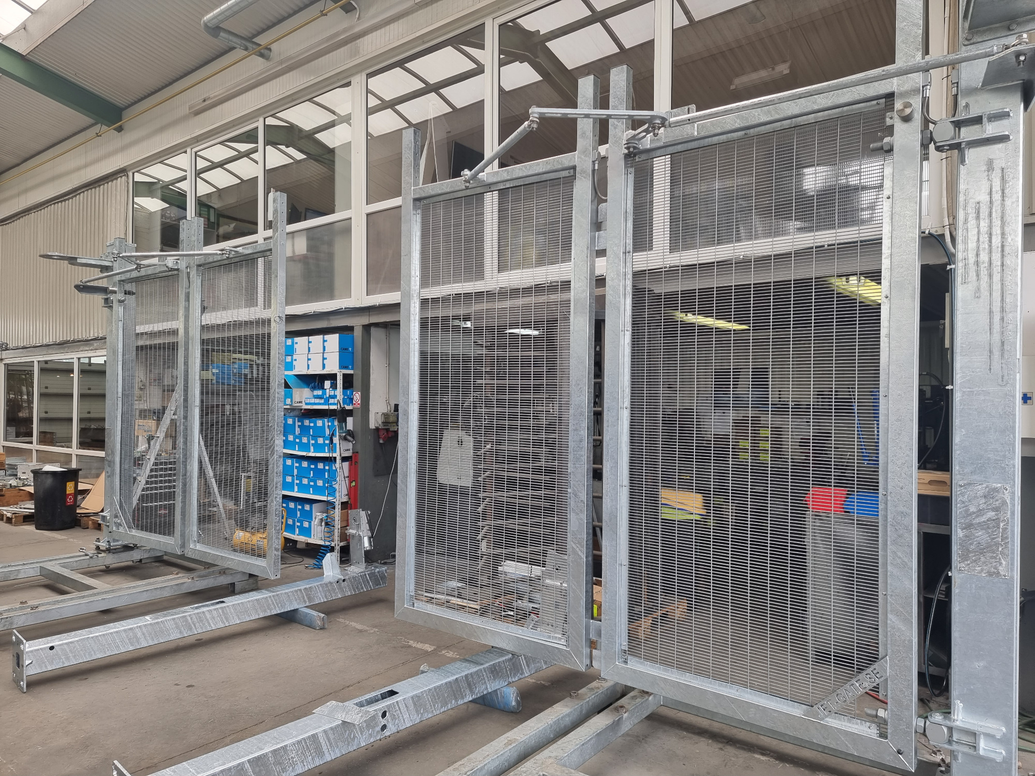 Siatkowe, metalowe segmenty bram przemysłowych w trakcie produkcji lub montażu w hali; widoczne elementy konstrukcyjne i akcesoria montażowe.