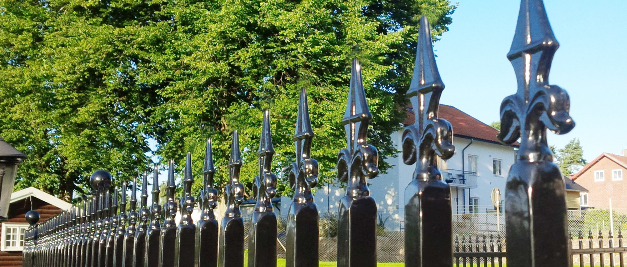 Szczegółowe ujęcie czarnego, metalowego ogrodzenia z dekoracyjnymi grotami w kształcie lilii, na tle zieleni drzew i jasnego budynku mieszkalnego.