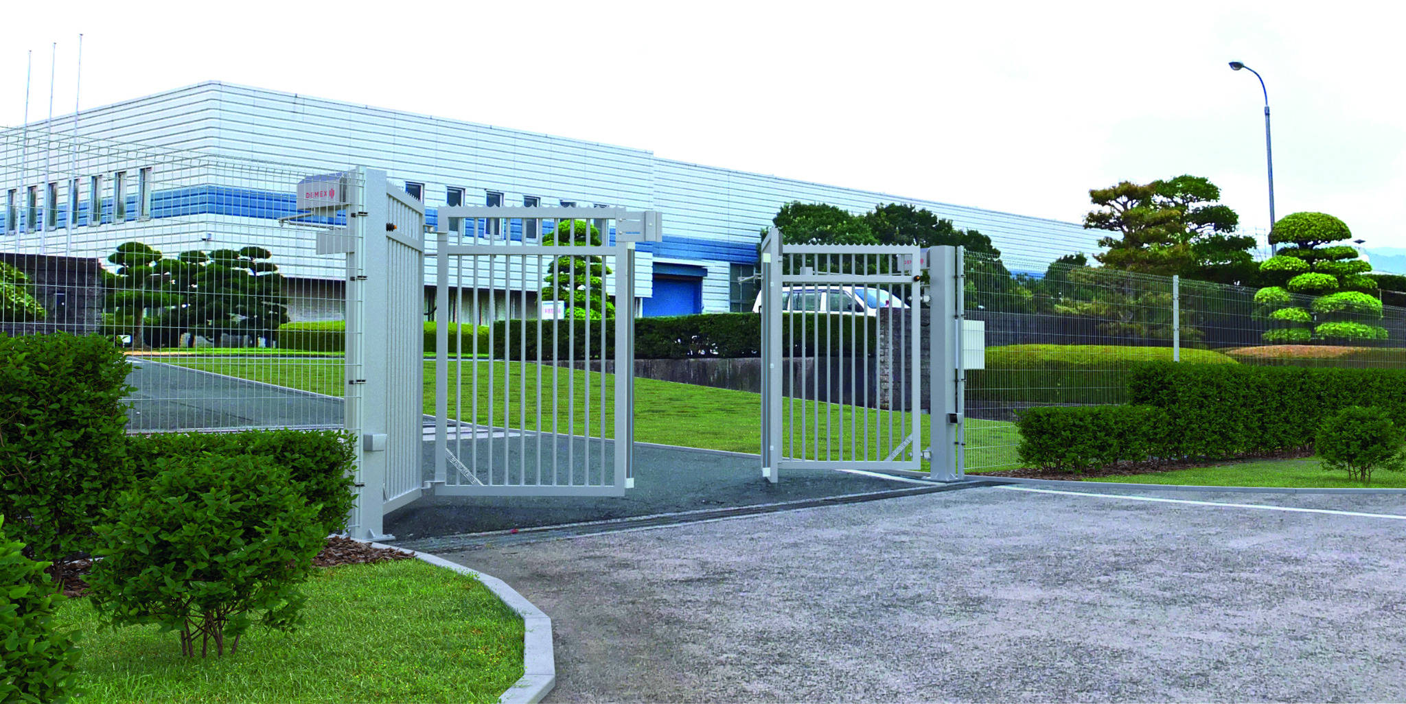 Otwarta, metalowa brama przemysłowa w kolorze szarym, z widocznym budynkiem przemysłowym w tle i zadbaną zielenią wokół.