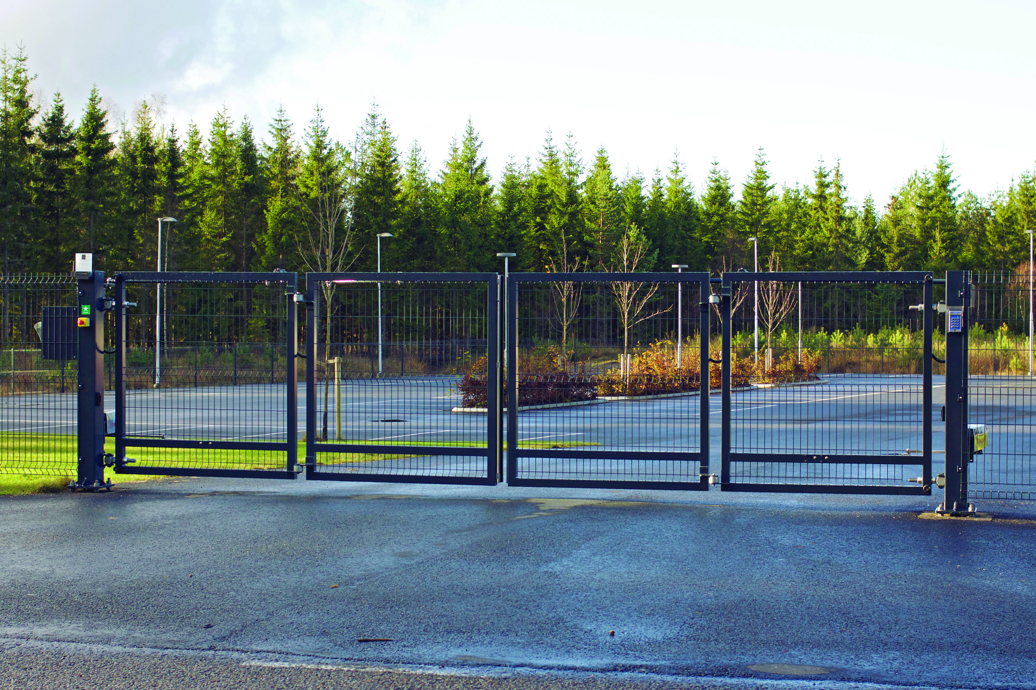 Czarna, czteroskrzydłowa brama przemysłowa z automatyką i panelem numerycznym, zamontowana na tle ogrodzenia z siatki i rzędu drzew iglastych.