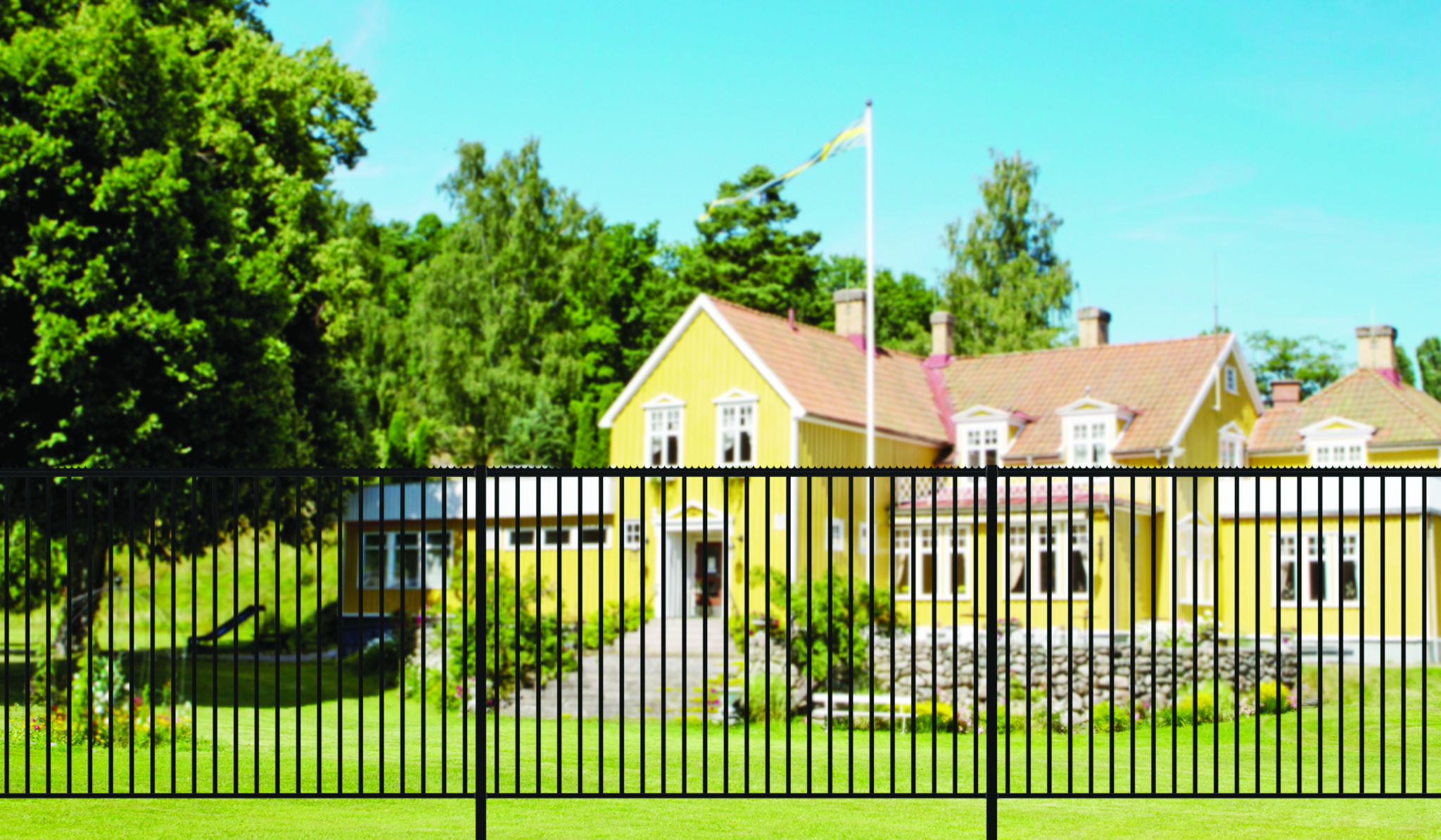 Nowoczesne, czarne ogrodzenie metalowe na tle rozmytego, żółtego domu z czerwoną dachówką i zielonym ogrodem w słoneczny dzień.