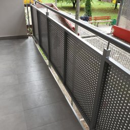 Eurobudowa - Szara, perforowana balustrada balkonowa z metalu na tle szarej podłogi z płytek, widok na zewnątrz z rusztowaniami w tle i elementami krajobrazu.