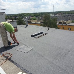 Eurobudowa - Prace dekarskie na dachu budynku: dekarz z palnikiem w ręku, butla gazowa obok, w tle zabudowa miejska i zieleń.