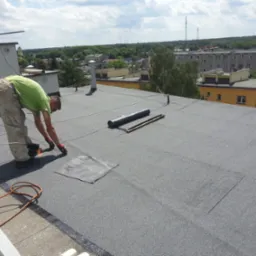 Prace dekarskie na dachu budynku: dekarz z palnikiem w ręku, butla gazowa obok, w tle zabudowa miejska i zieleń.