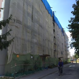 Eurobudowa - Wielopiętrowy budynek w trakcie ocieplania, osłonięty siatką z widocznym logo Weber Terranova, z niebieskim zabezpieczeniem na dachu, na pierwszym planie rowerzysta na ulicy.