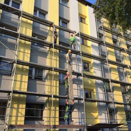 Eurobudowa - Pracownicy na rusztowaniu montują elementy elewacji budynku w odcieniach żółci i szarości.