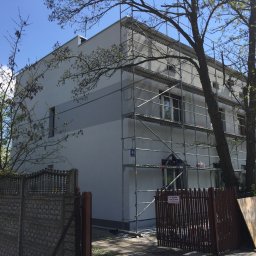 Eurobudowa - Dwukondygnacyjny dom w trakcie malowania elewacji, otoczony rusztowaniem, widoczny fragment ogrodzenia i drzewa.