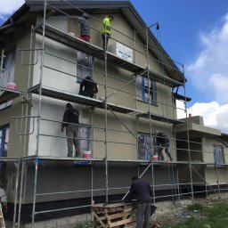 Eurobudowa - Ekipa budowlana na rusztowaniu podczas nakładania tynku na elewację domu jednorodzinnego, okna zabezpieczone folią, widoczne puszki z farbą i paleta z materiałami.