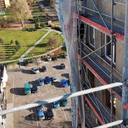 Eurobudowa - Prace na wysokościach: Robotnicy na rusztowaniu przy ocieplaniu bloku mieszkalnego, widok z góry na parking i park.
