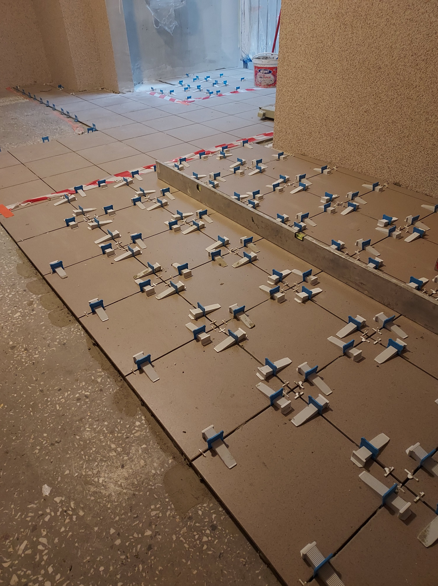 Proces układania płytek podłogowych z użyciem systemu poziomowania, widoczna poziomica i wiadro z zaprawą w tle.