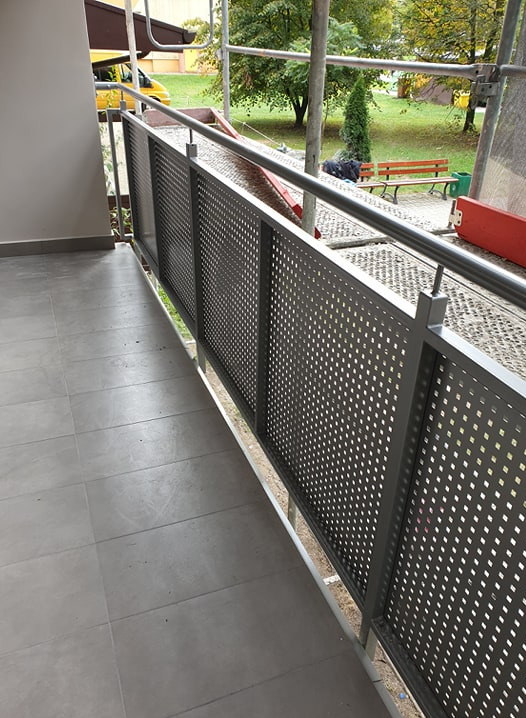 Szara, perforowana balustrada balkonowa z metalu na tle szarej podłogi z płytek, widok na zewnątrz z rusztowaniami w tle i elementami krajobrazu.
