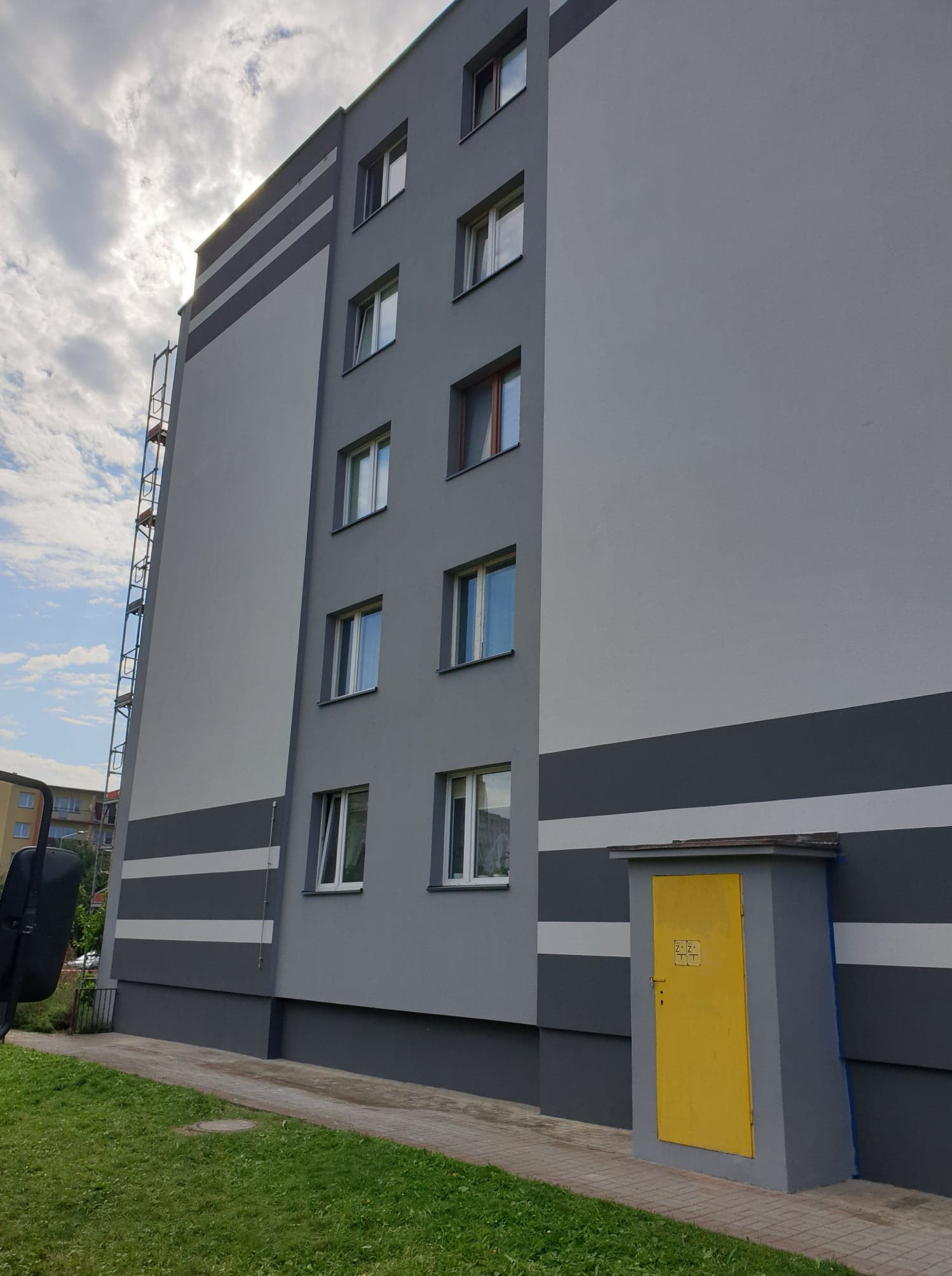 Wielopiętrowy budynek po remoncie elewacji w odcieniach szarości z poziomymi pasami i żółtą szafką przy chodniku, widoczne rusztowanie przy ścianie.