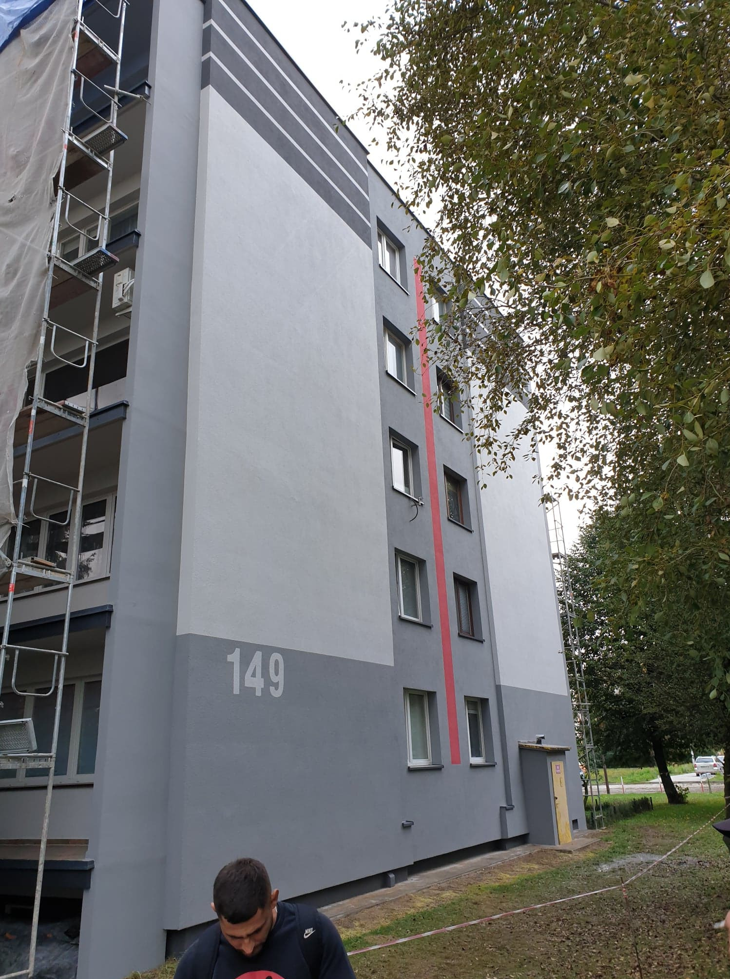 Wielopiętrowy budynek po odnowieniu elewacji w odcieniach szarości, z numerem 149 i pionowym czerwonym pasem, widoczne rusztowania i fragment osoby w dolnej części kadru.