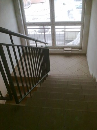 Klatka schodowa z metalową barierką, widok z góry na schody pokryte płytkami ceramicznymi, duże okno z widokiem na zewnątrz, częściowo zasłonięte folią ochronną.