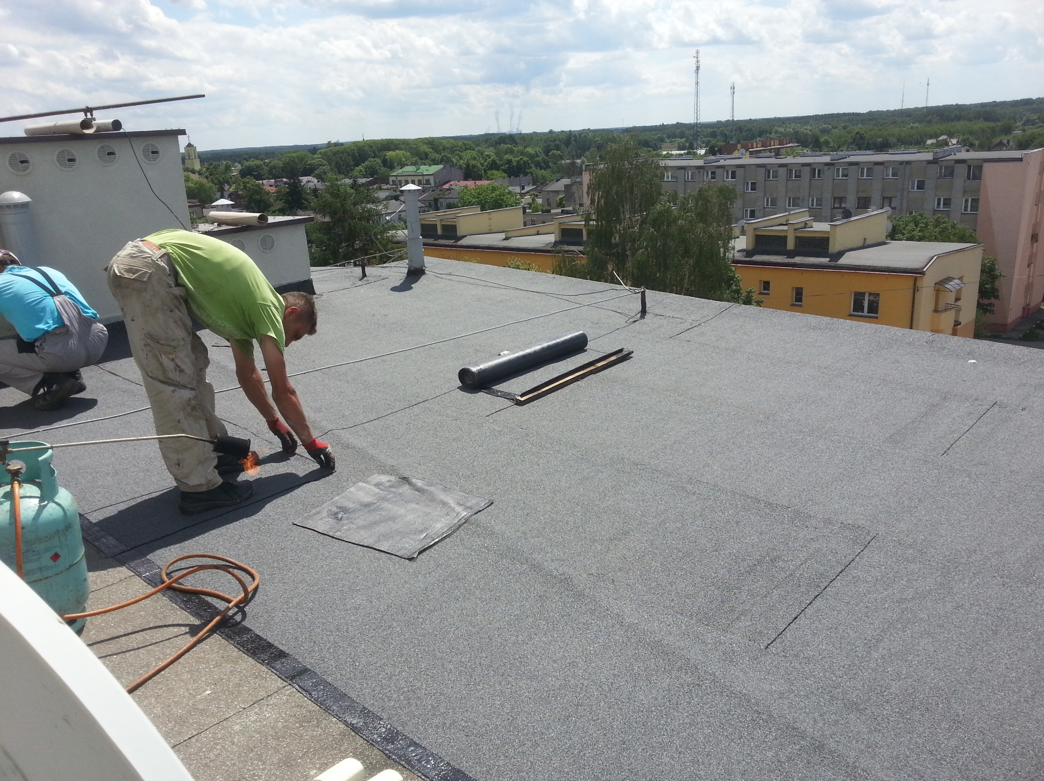 Prace dekarskie na dachu budynku: dekarz z palnikiem w ręku, butla gazowa obok, w tle zabudowa miejska i zieleń.